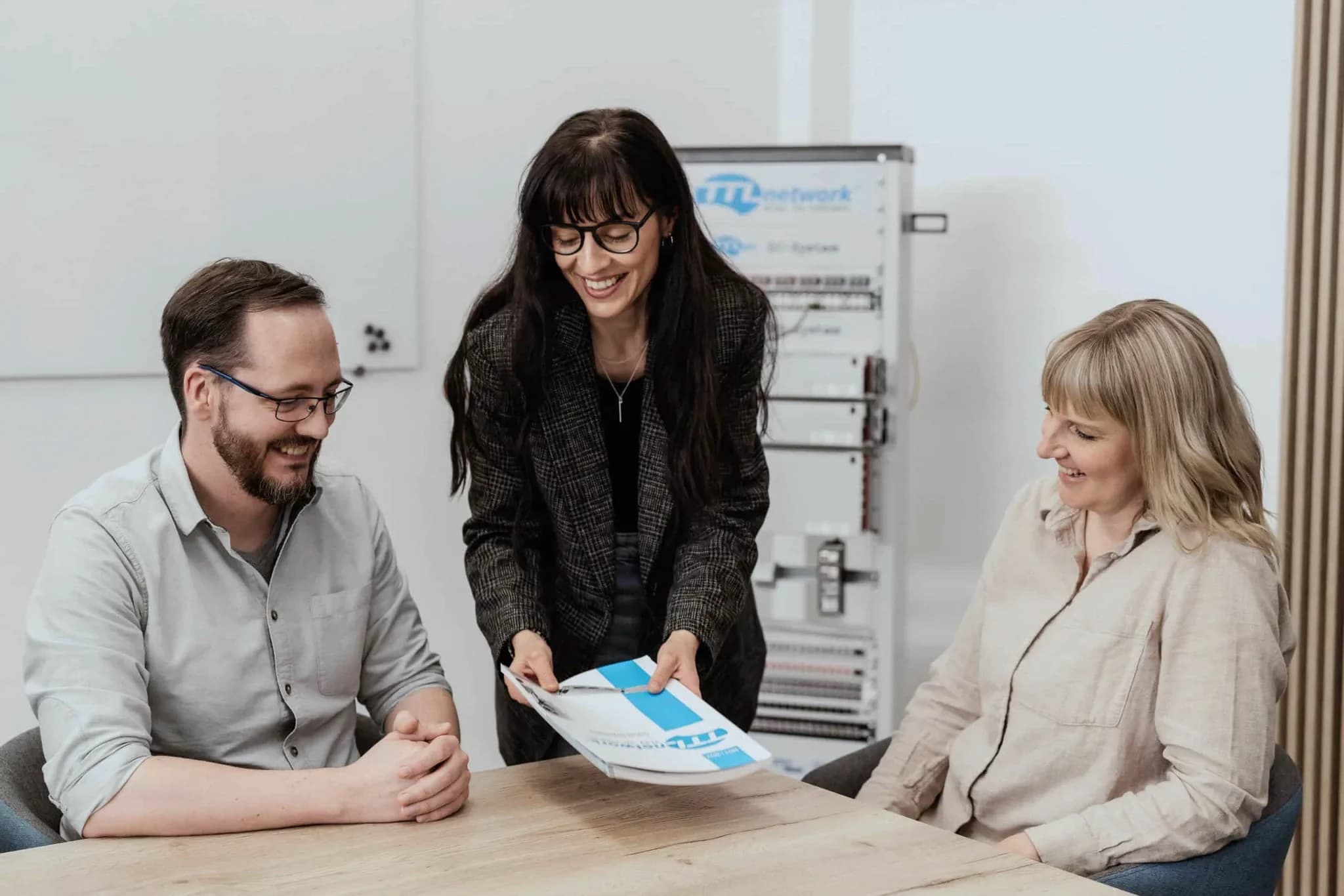 A TTL Network employee shows two guests a brochure at the meeting table, with a network cabinet bearing the TTL logo behind them
